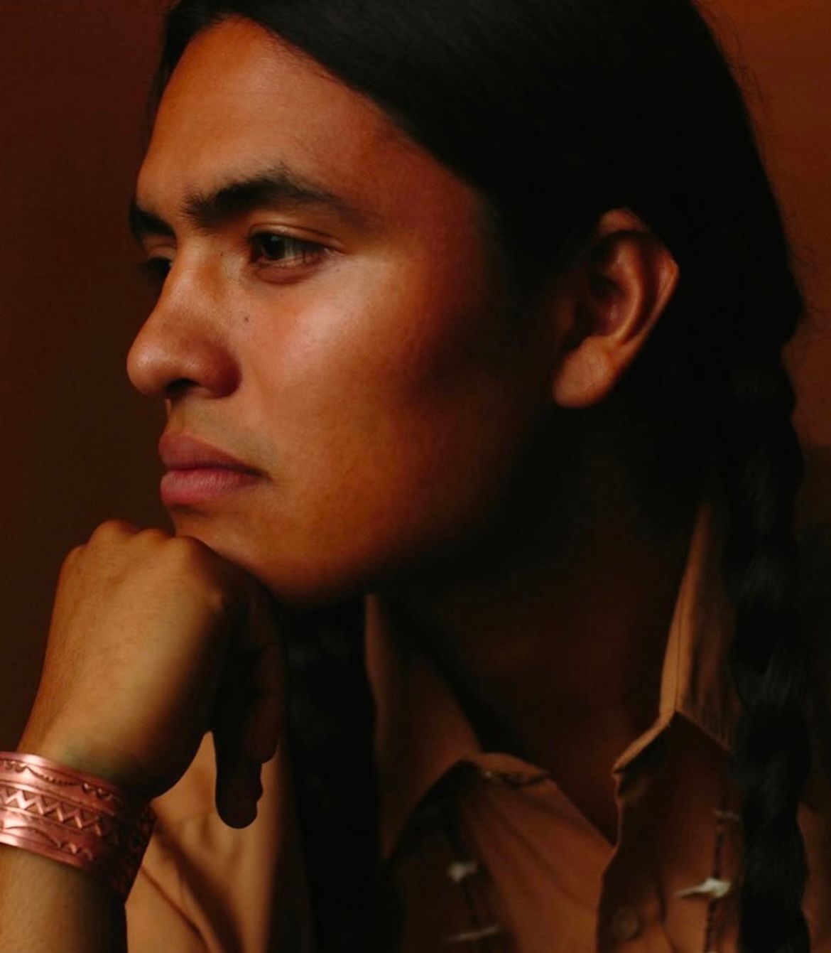 Man with long dark hair in a side profile, wearing a copper bracelet, looking pensive.