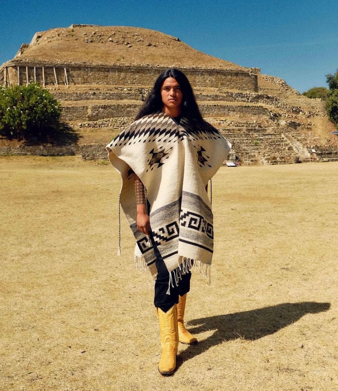 Woman in poncho and yellow boots stands before ancient pyramid ruins.