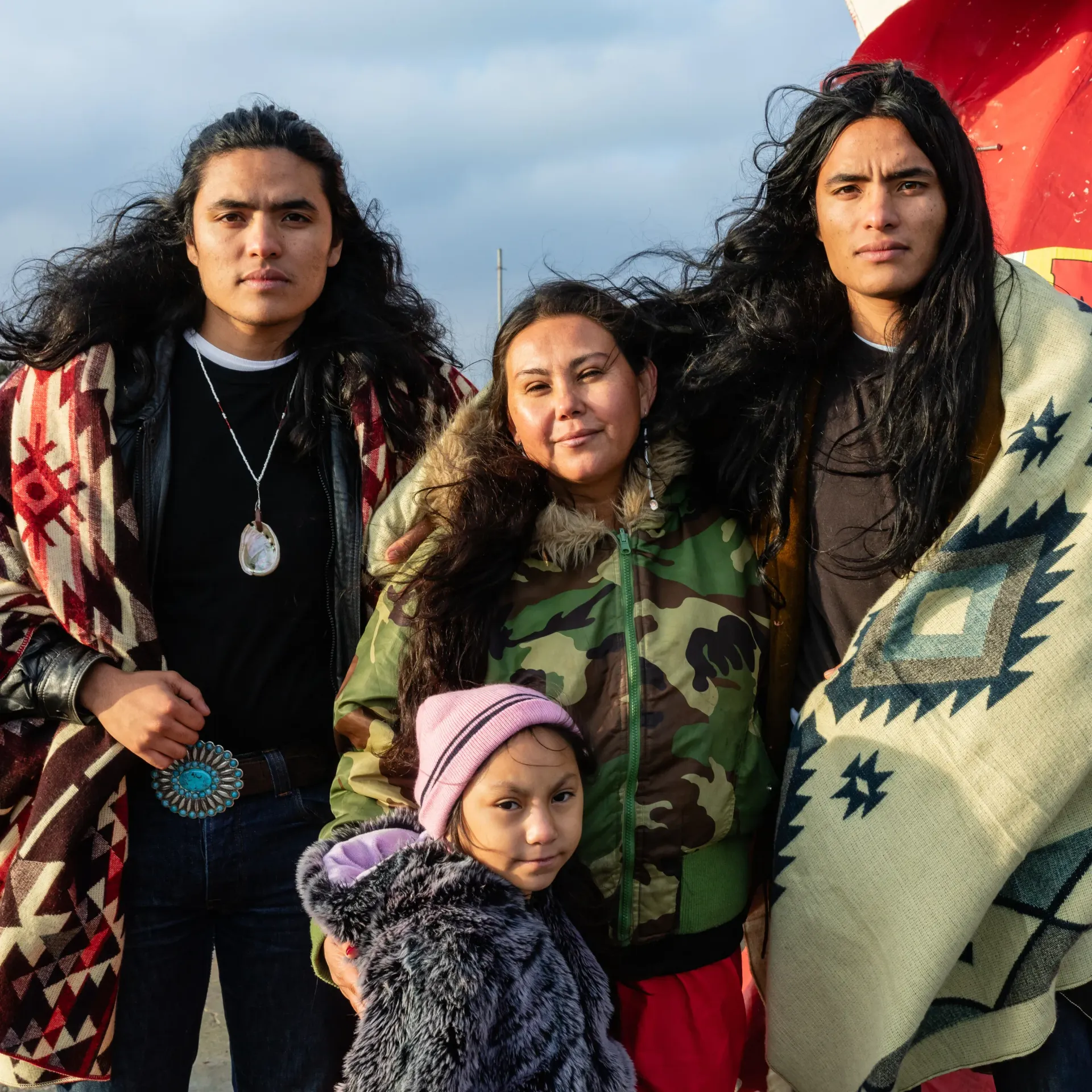 Family of four bundled in outerwear, standing outdoors. Two men with long hair flank a woman and child.
