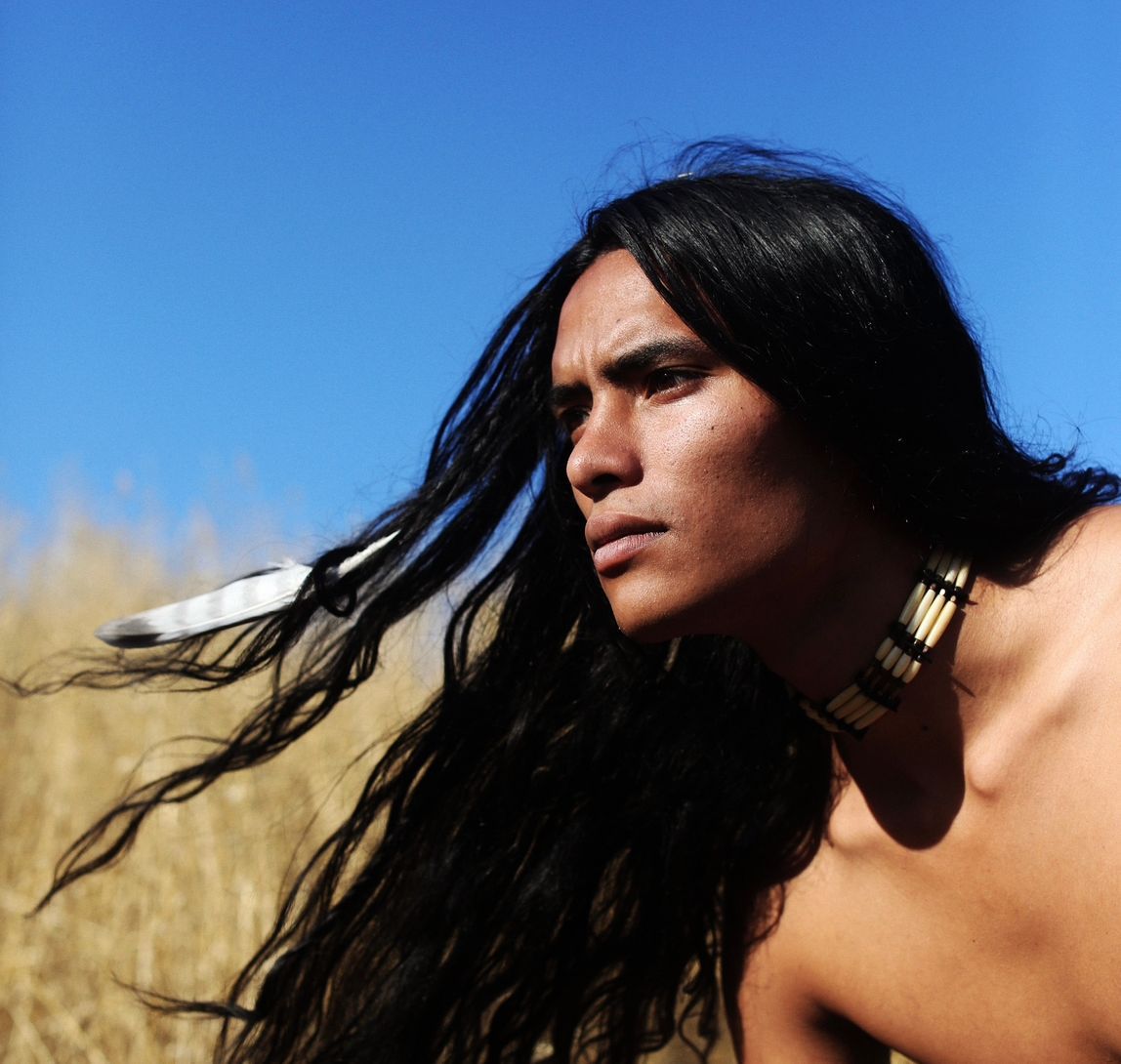 Man with long, dark hair and feather in hair, looking toward the right. Blue sky and dry grass in the background.