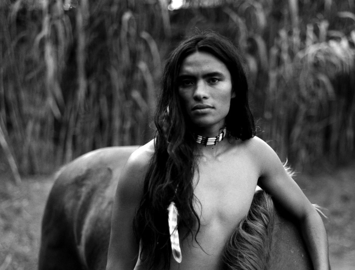 Person with long dark hair, bare-chested, wearing a necklace and feather, leans on a horse.