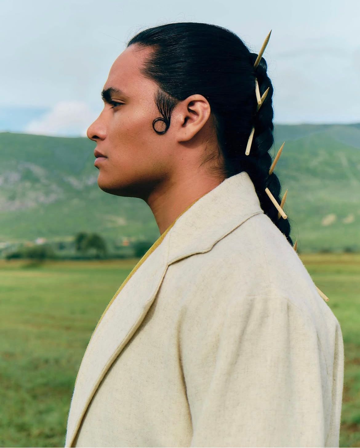 Person in cream jacket with braided hair adorned with gold pins, outdoors with green field and hills.