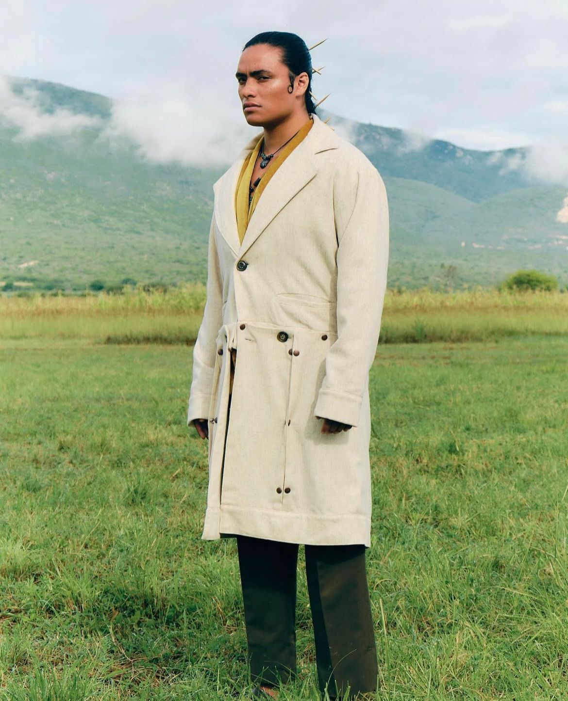 Person in long cream coat stands in a grassy field with mountains in the background.