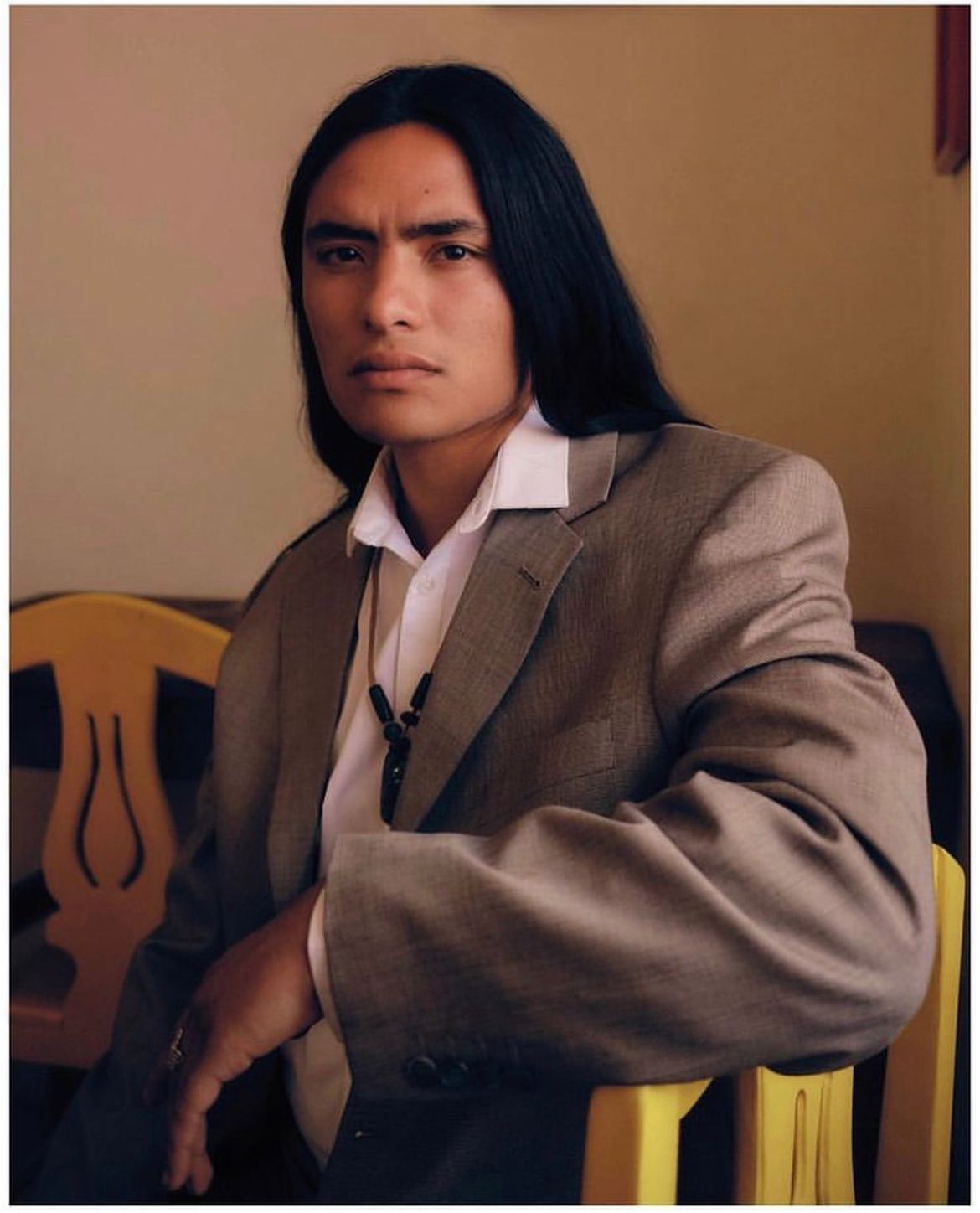 Man with long black hair, in a blazer and white shirt, sitting with a serious expression, arms resting on a yellow chair.