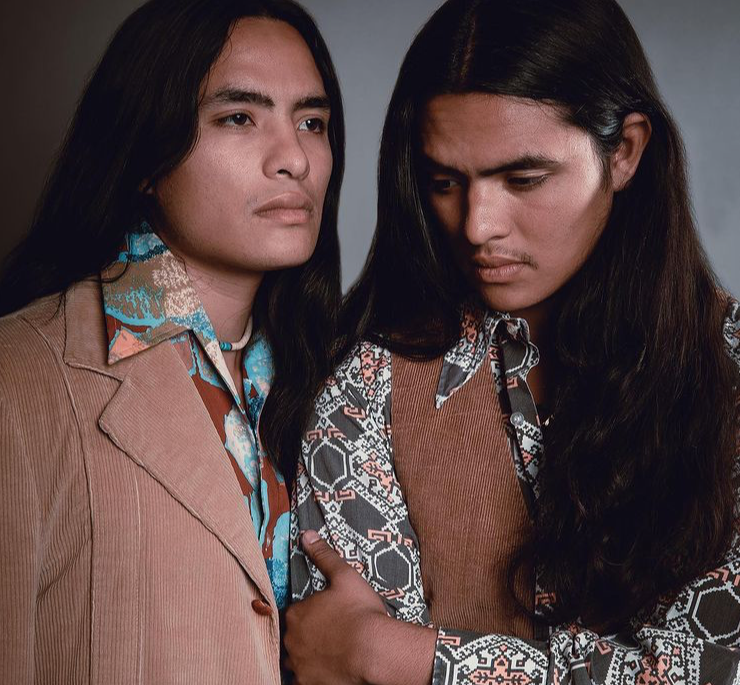 Two men with long dark hair, wearing patterned shirts and a corduroy jacket, looking serious.