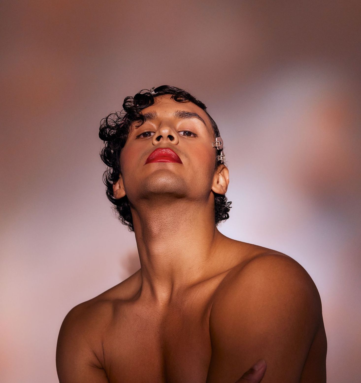 Person with red lipstick, looking up, shirtless. Curly hair, pink backdrop.