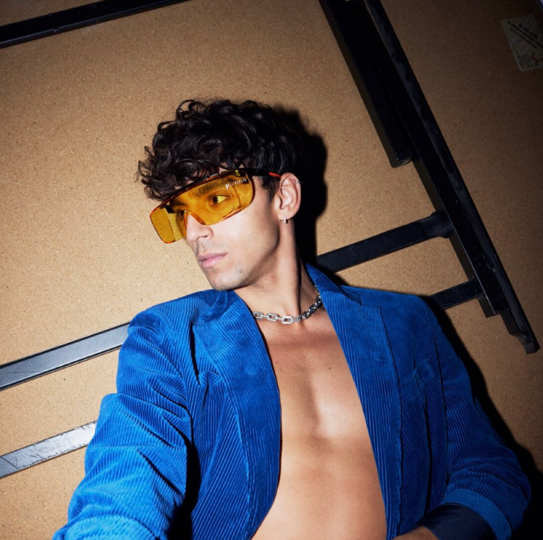 Man with curly hair and yellow sunglasses, blue blazer, silver chain, looking away from camera.