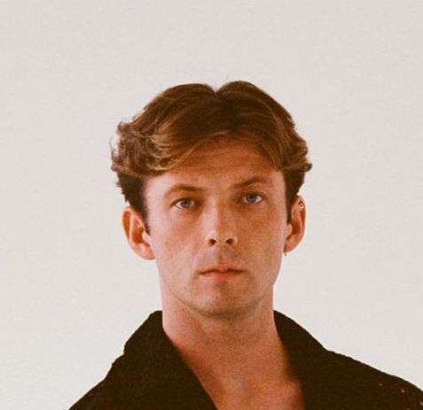 Man with light brown hair looking forward, wearing a dark textured jacket, against a white background.