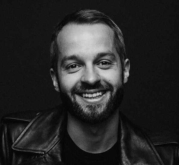 Man with a beard smiling, wearing a leather jacket. Black and white, studio shot.