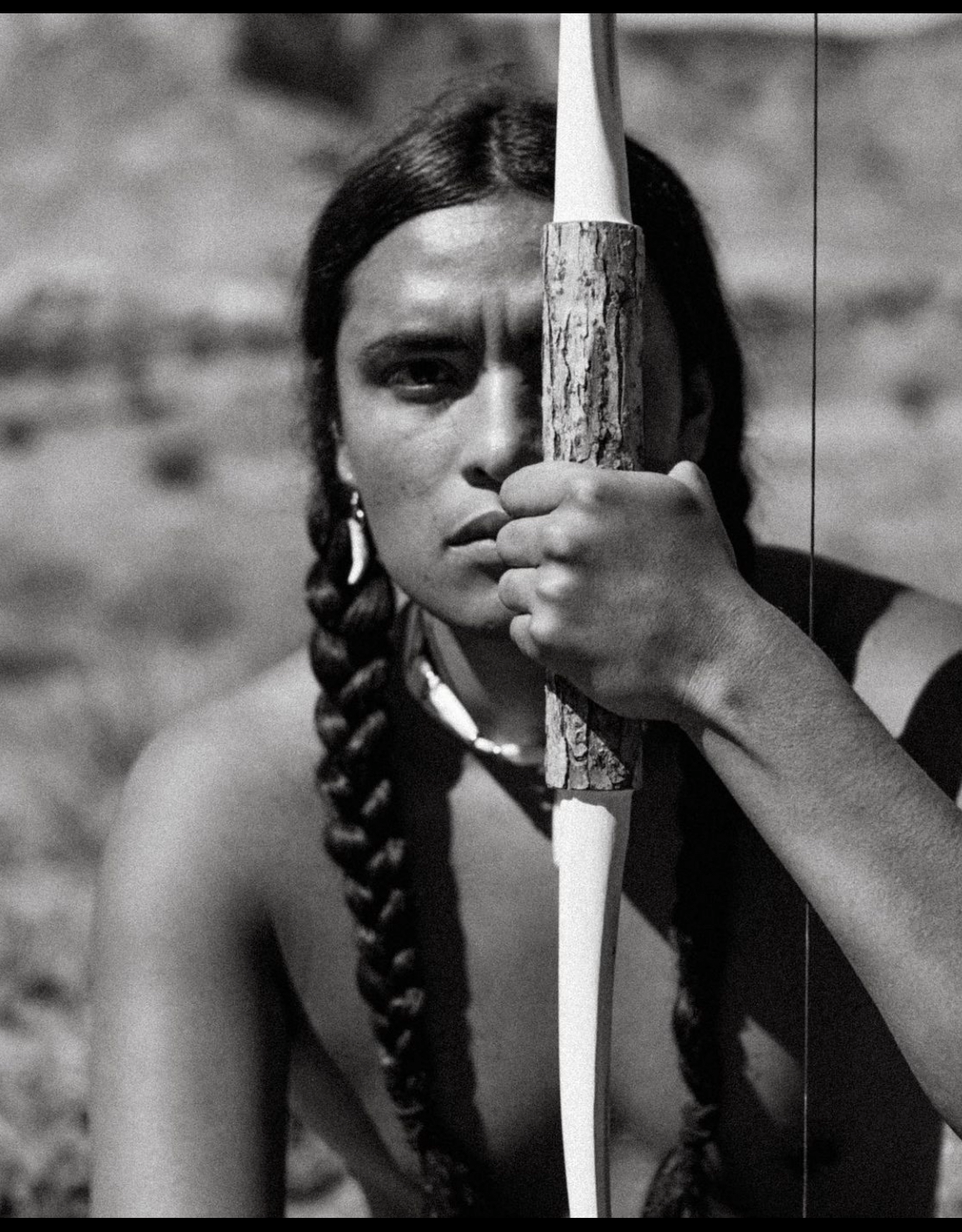 Person holding a bow, looking directly at the viewer. One braided pigtail visible, with necklace.
