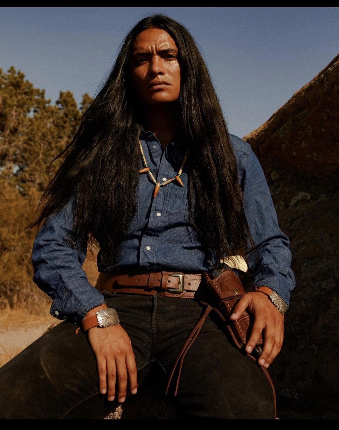 Man with long dark hair, wearing a blue shirt, belt, and holster, sitting outdoors, looking serious.