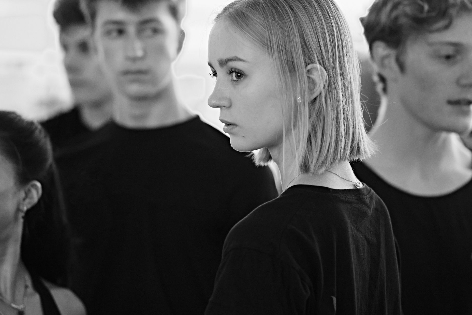 Woman with crimped hair in a group of people, possibly dancers, wearing black shirts, looking off-center.