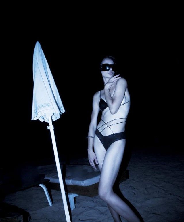 Woman in a black swimsuit and eye mask stands on a beach next to a closed umbrella.