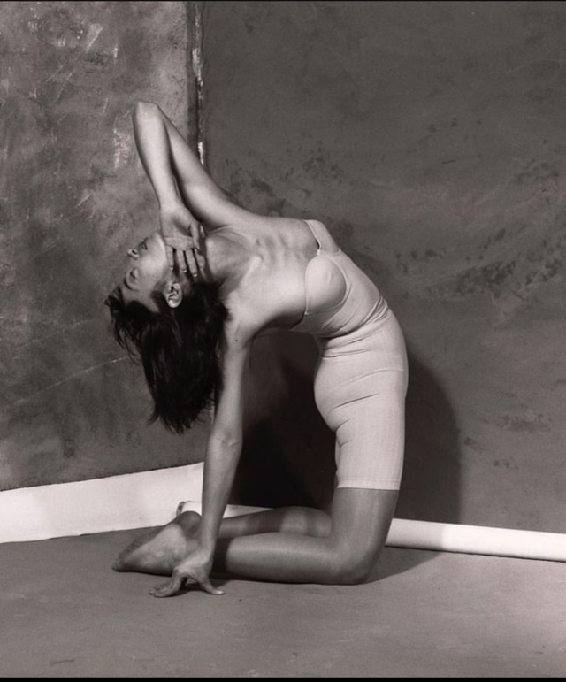 Woman in yoga pose, arched back, knees bent, arms raised, against a wall.