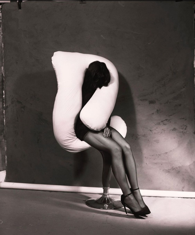 A woman wearing black lingerie and stockings is contorted in a white, sculptural chair.