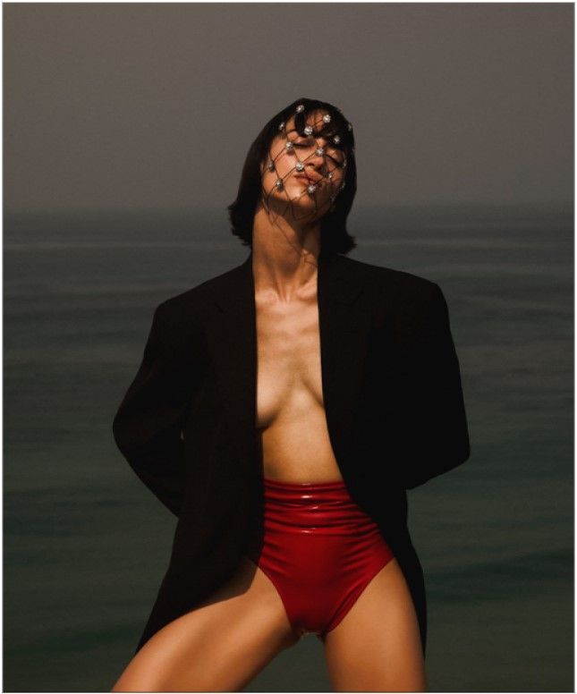 Woman in black blazer and red high-waisted shorts, facing away from the camera, with ocean in the background.