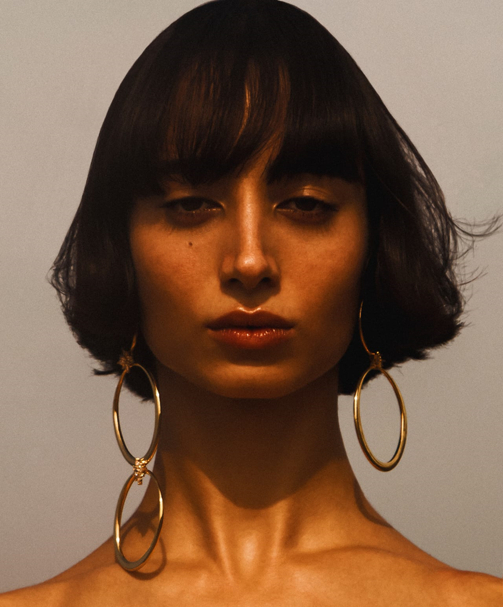 Woman with short dark hair and bangs, wearing large gold hoop earrings, looking directly at the viewer.