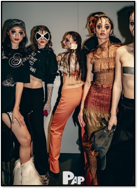 Models backstage at a fashion show: posing in various outfits with elaborate makeup, against a plain background.