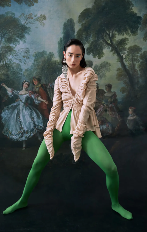 Person wearing a light-colored top and bright green tights poses in front of a classical painting.