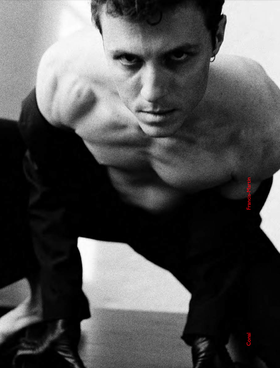 Man in dark suit, chest bare, leans forward intensely. Black and white.