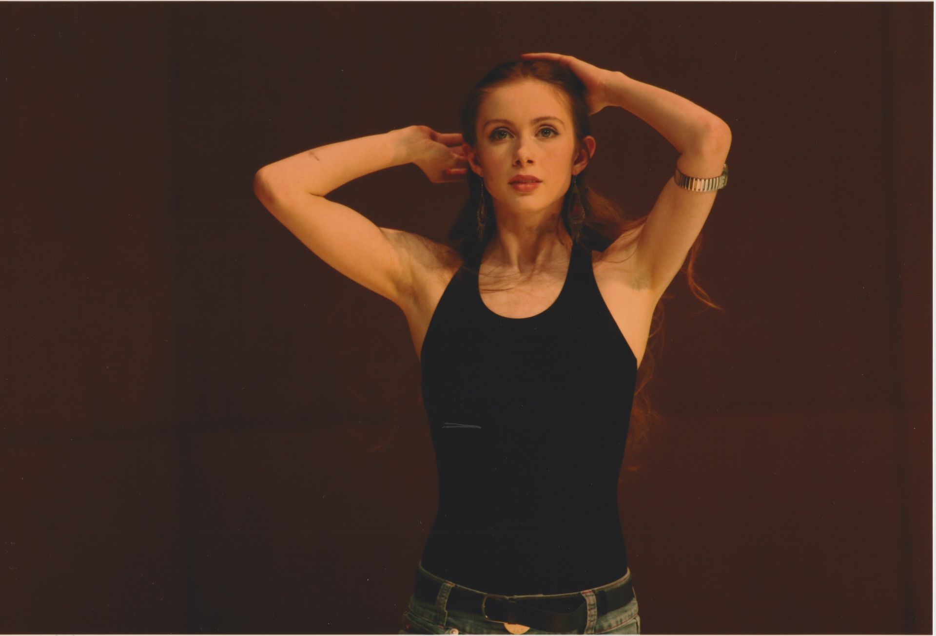 Woman in black tank top with hands in hair against a dark backdrop.