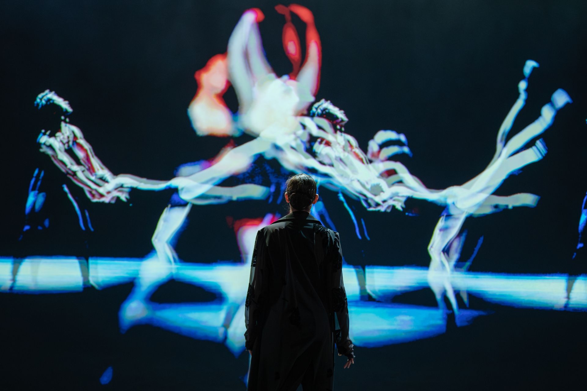 A person stands before a projection of dancers. The projection shows multiple figures with blurred, colorful lines.
