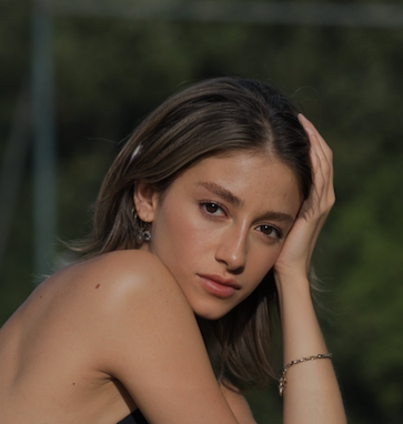 Woman with light brown hair, arm resting on head, looking towards camera. Outdoors, green background.