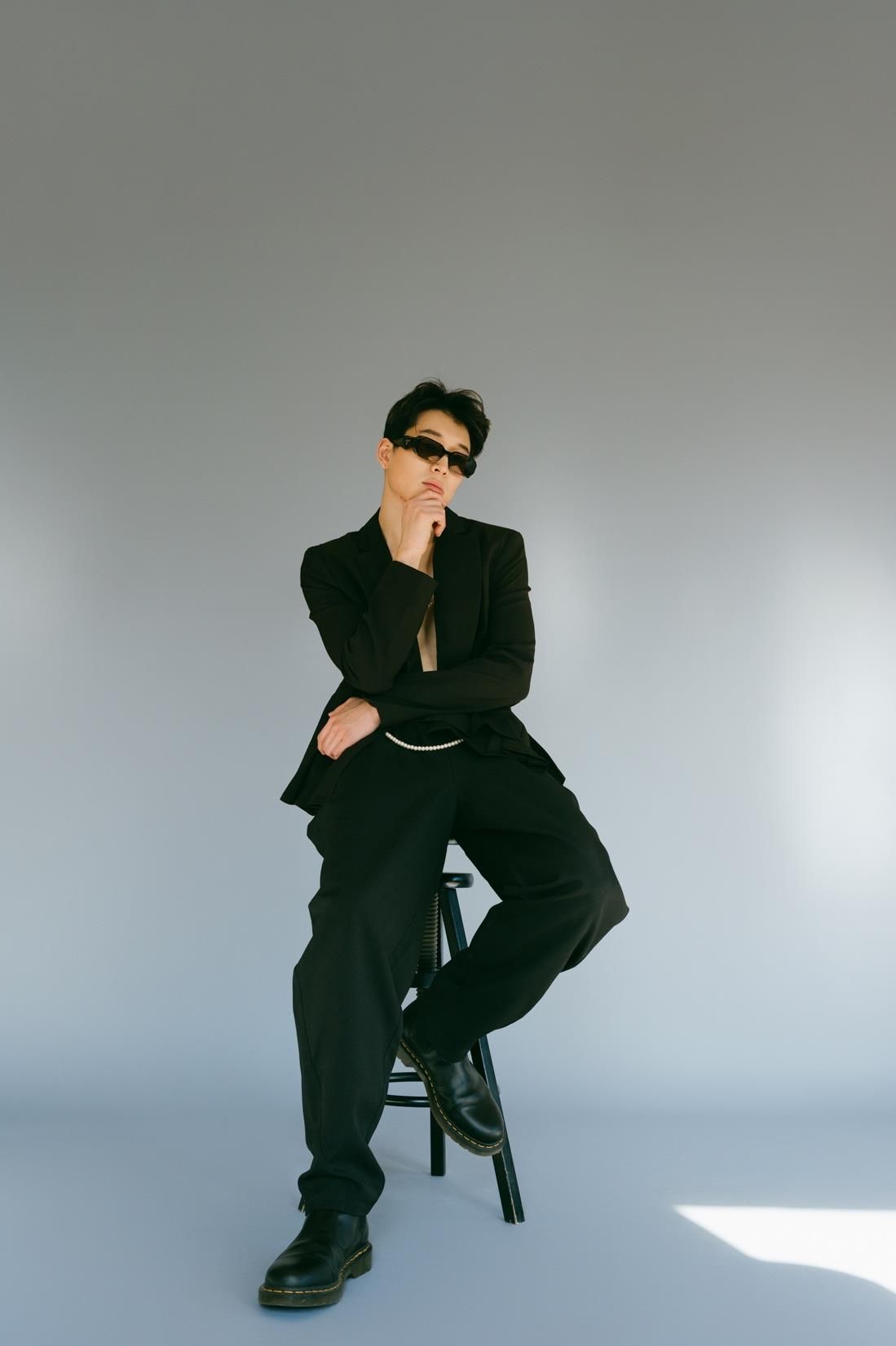 Person in black suit and sunglasses, sitting on a stool, leaning chin on hand, gray background.