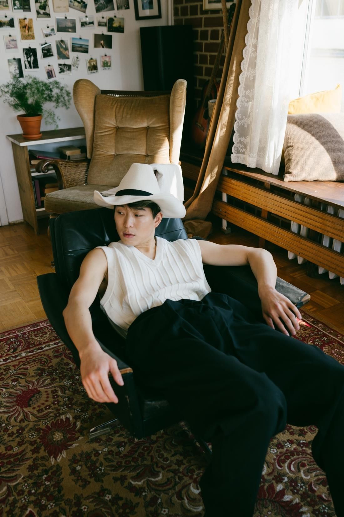 Person in white vest and cowboy hat lounges in a chair near a window and red rug.