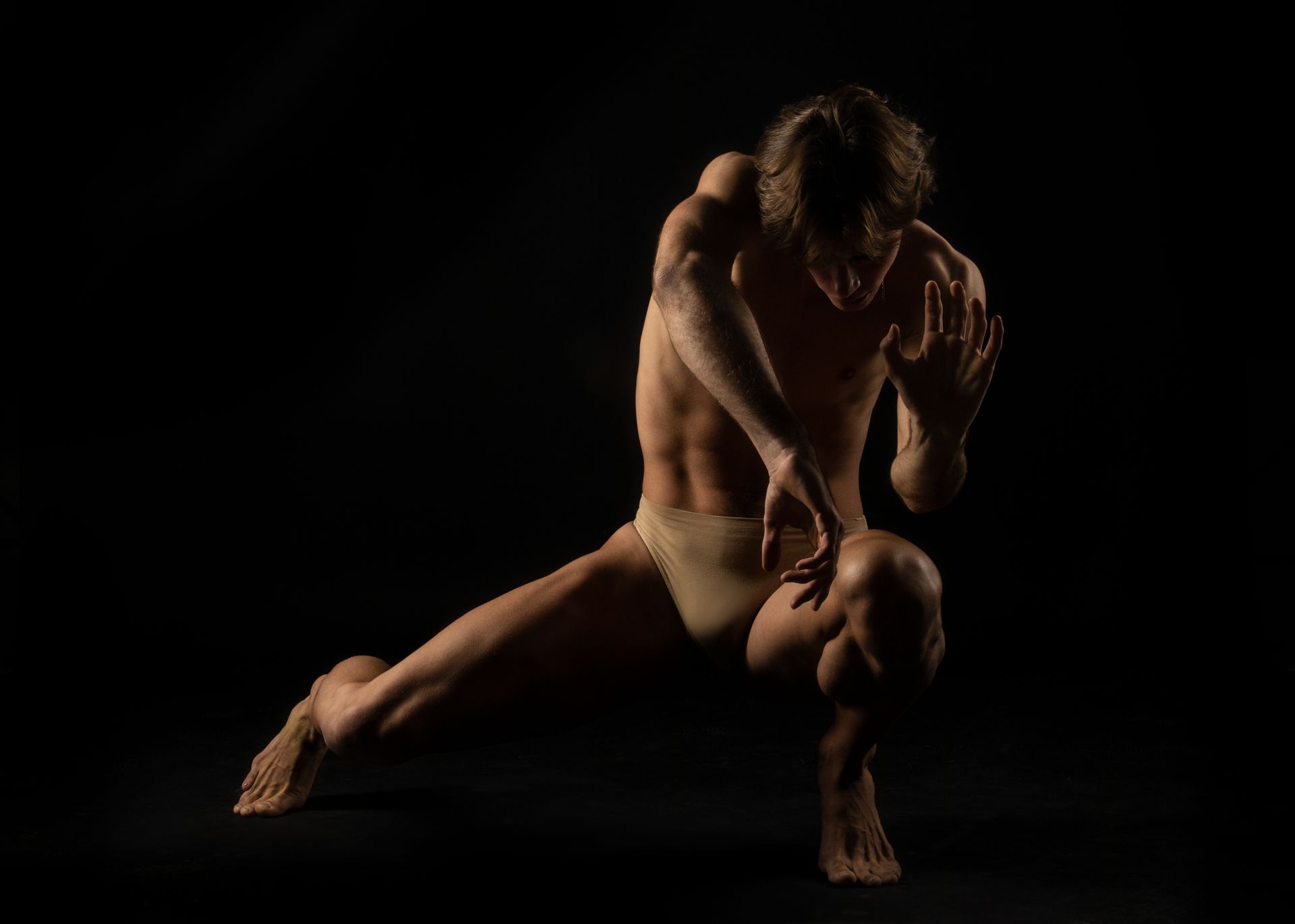 Male dancer in beige briefs, crouching low with arms outstretched against a black background.