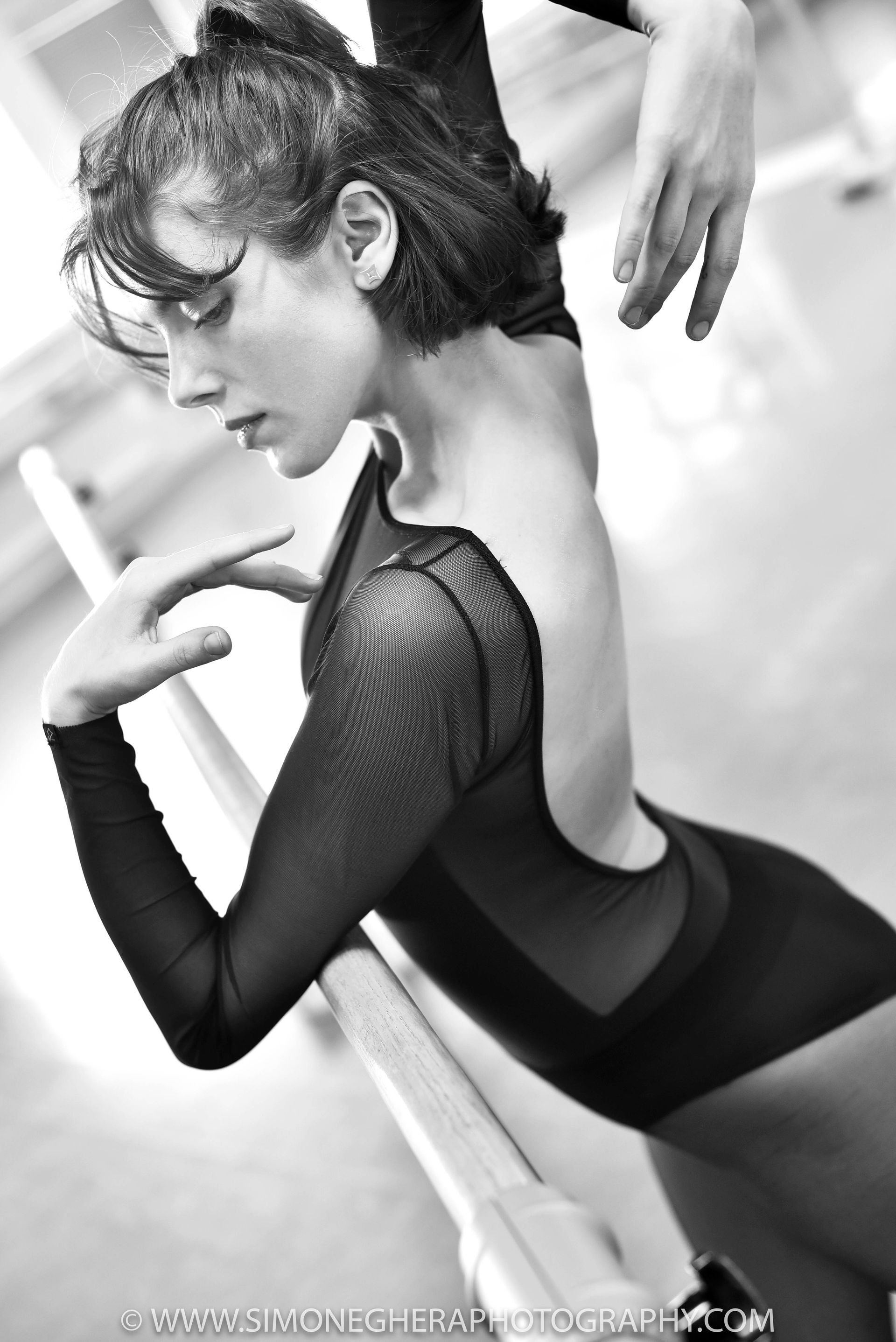 Ballet dancer in a black leotard leans against a barre. Black and white photo.