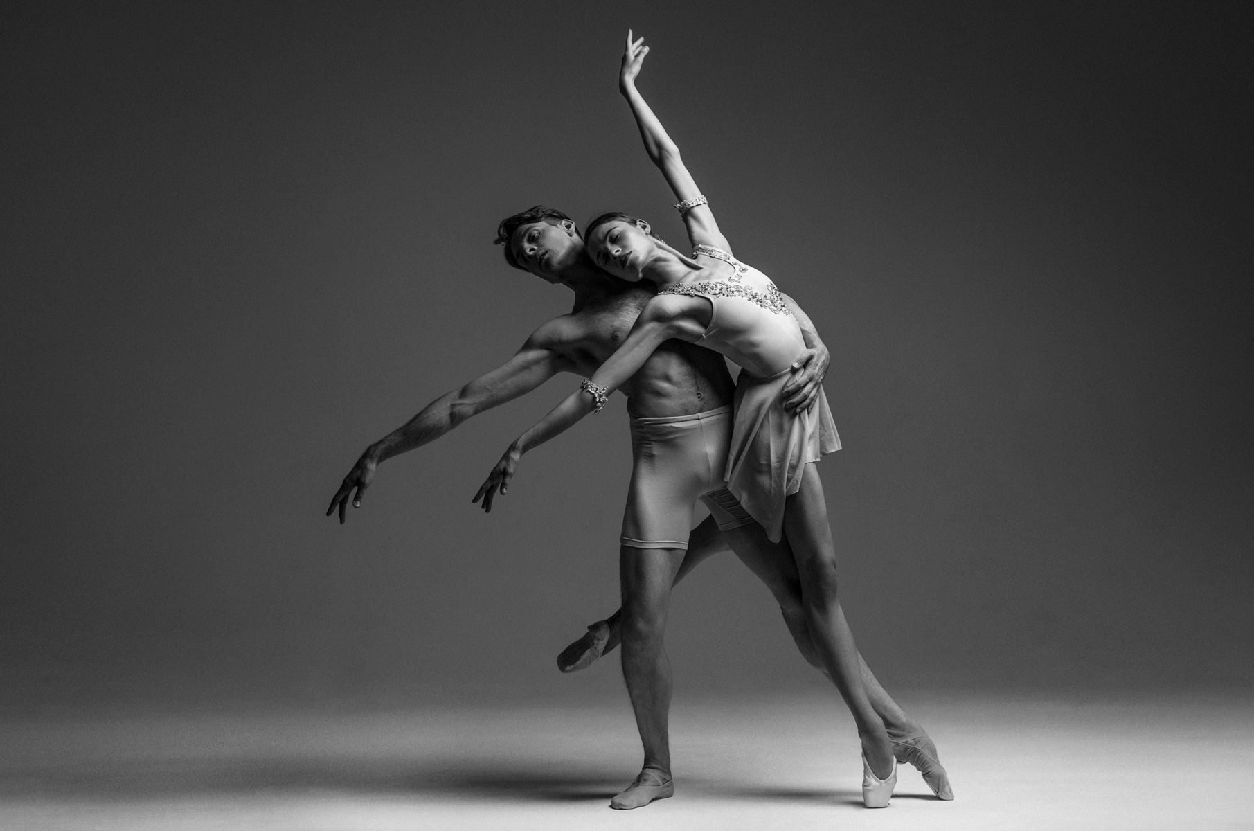 Two ballet dancers performing a graceful, partnered pose; man supports woman leaning back. Black and white.