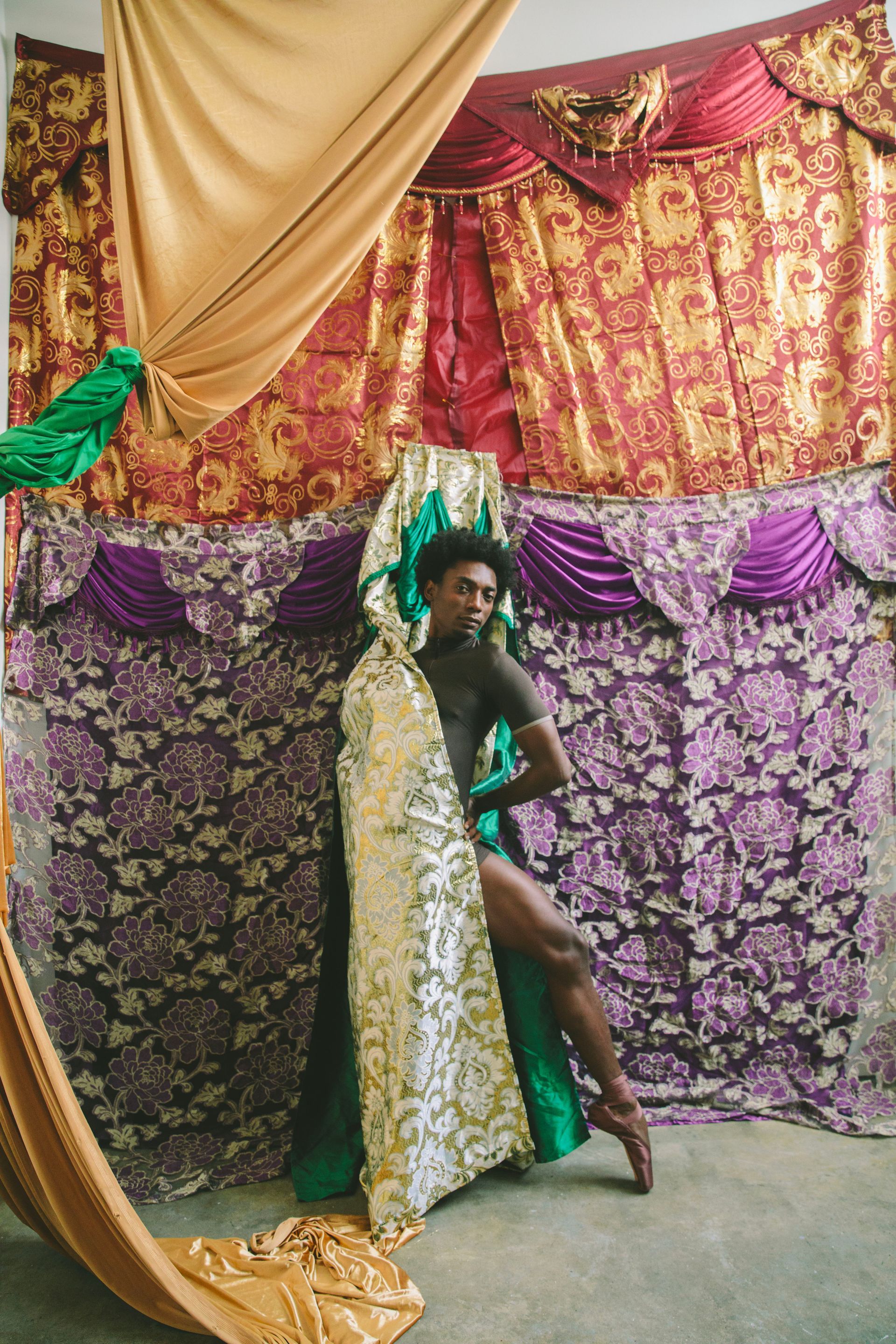 A person in a black leotard poses in front of ornate, patterned drapes.