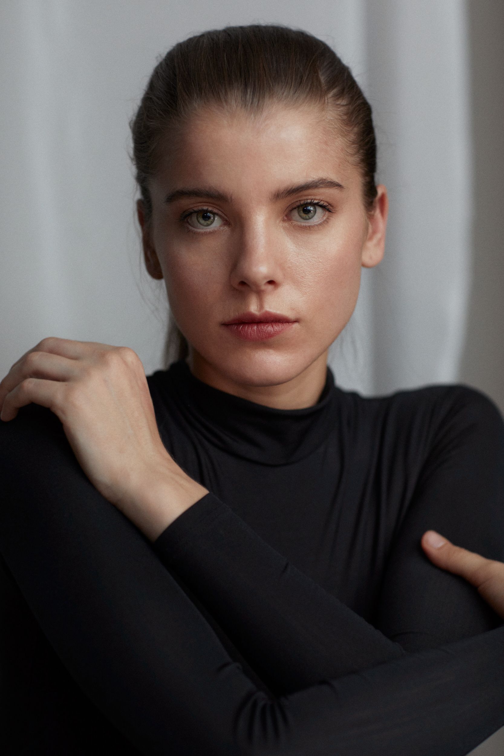 Woman with light eyes, dark hair in a ponytail, wearing a black turtleneck, arms crossed, neutral expression.