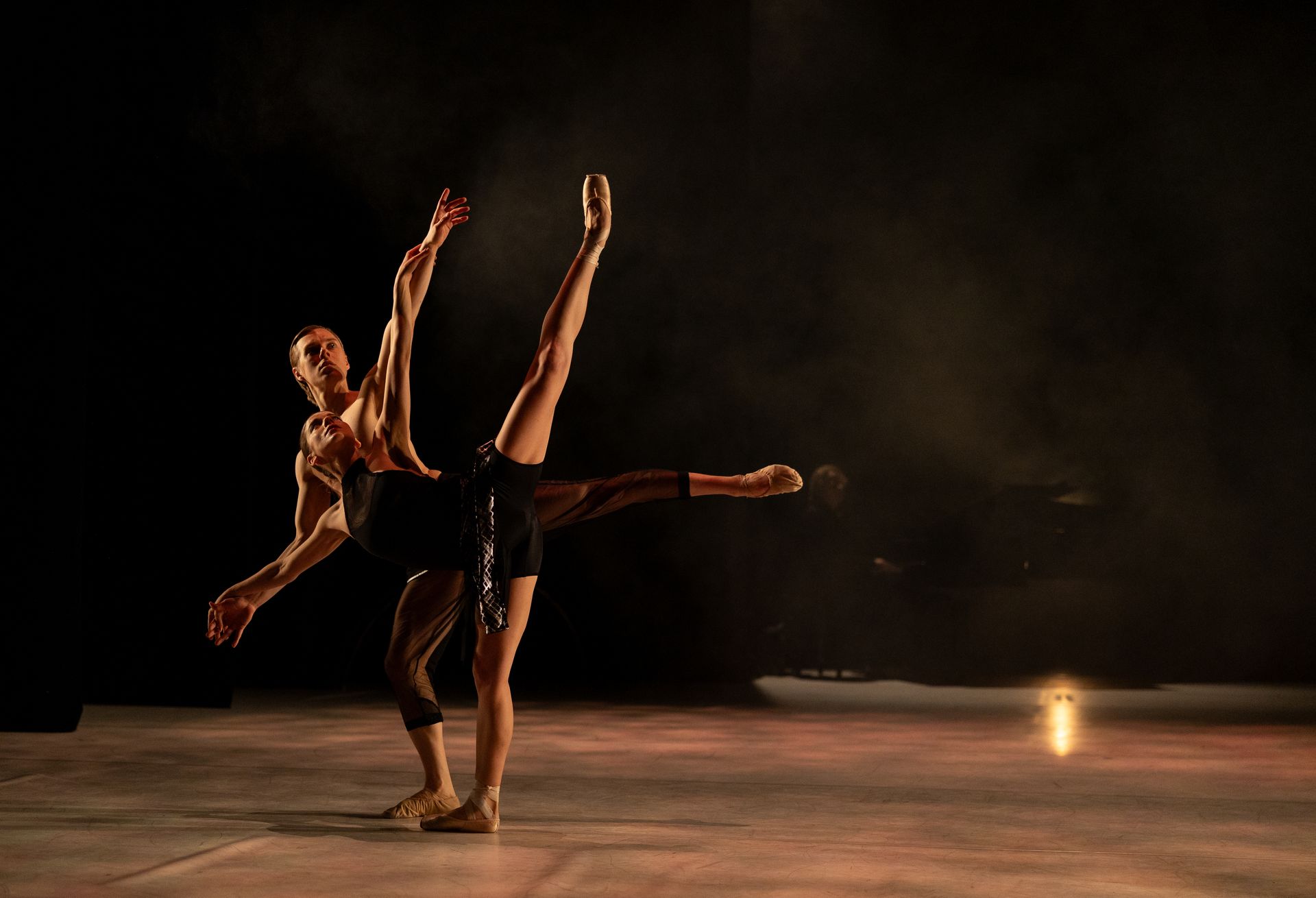 Two dancers on a stage, lit with a spotlight. One leg lifted high, arms extended. Dark clothing.