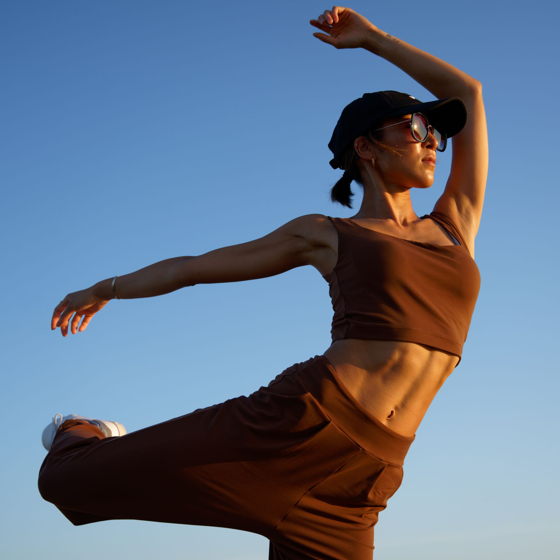 Woman in brown activewear, balancing on one leg, arm raised against a blue sky.