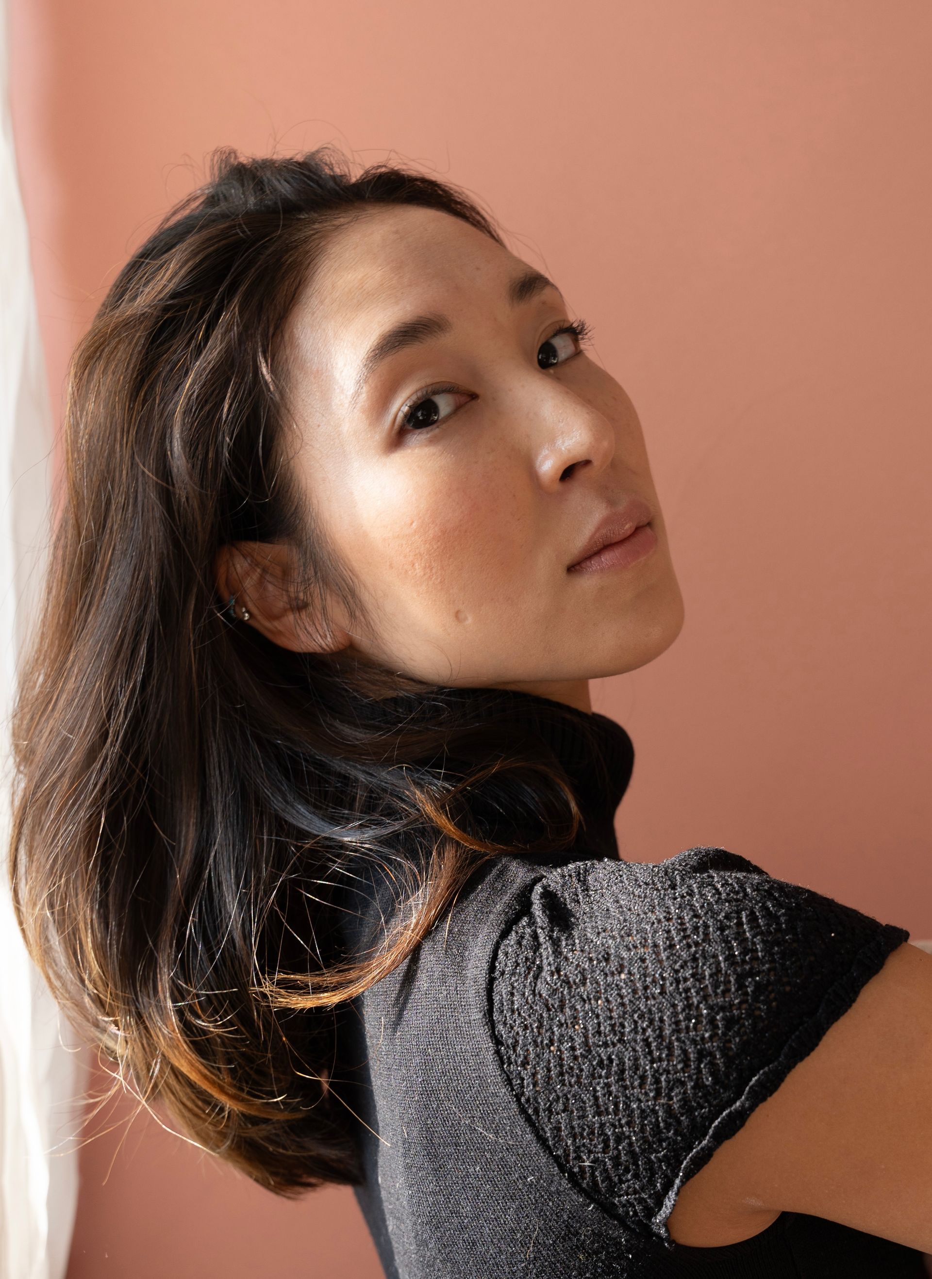 Woman with dark hair looking over her shoulder, wearing a black turtleneck. Pink background.