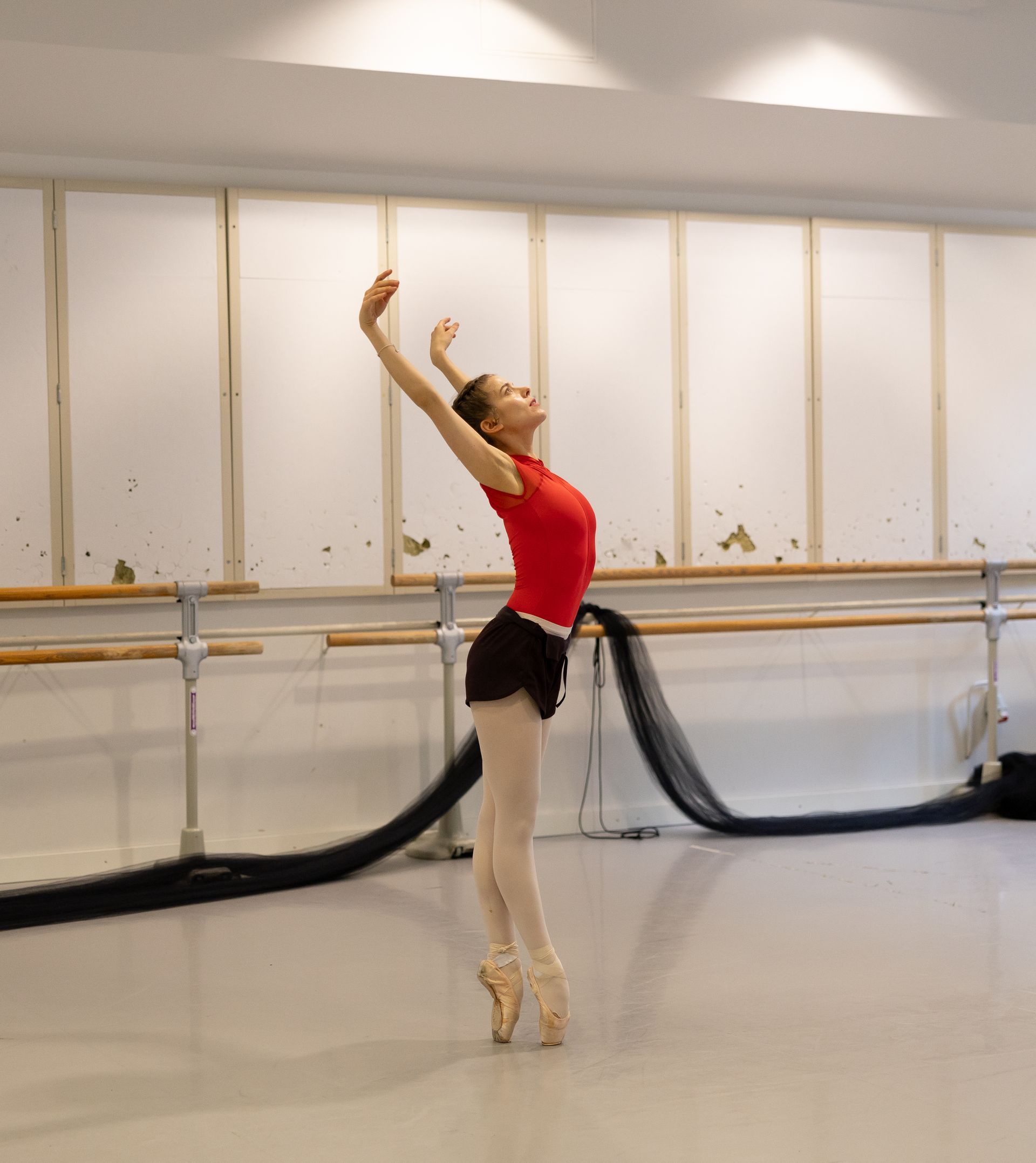 Ballet dancer en pointe, arms raised, in a studio with a barre. Wearing red top, black shorts, and pointe shoes.