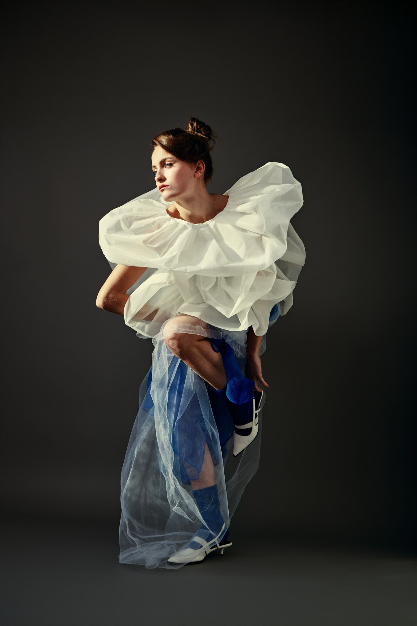 Woman in puffy white collar and blue dress squats, looking left on gray background.