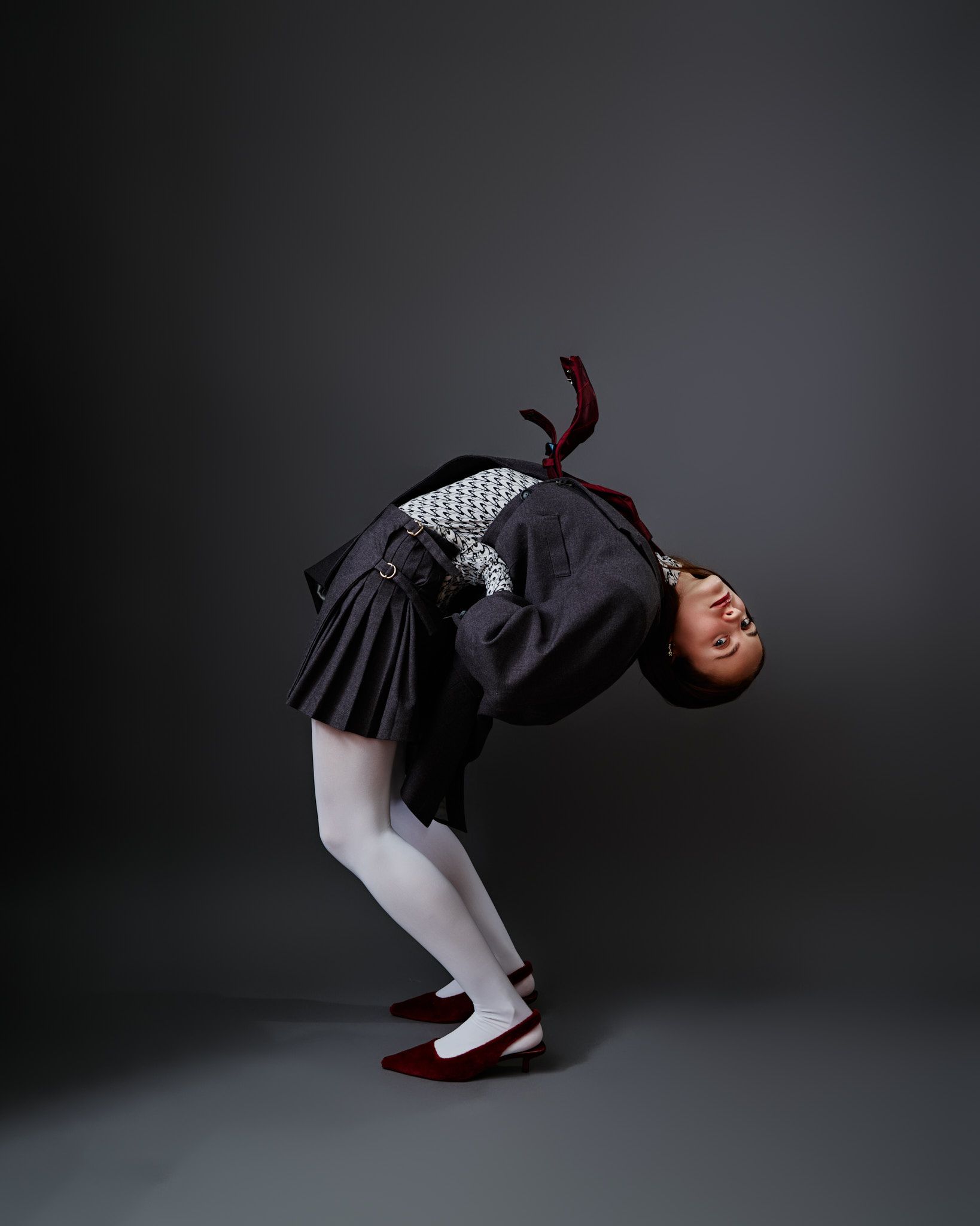 Person arching backwards, wearing patterned jacket, pleated skirt, white tights, and red shoes.
