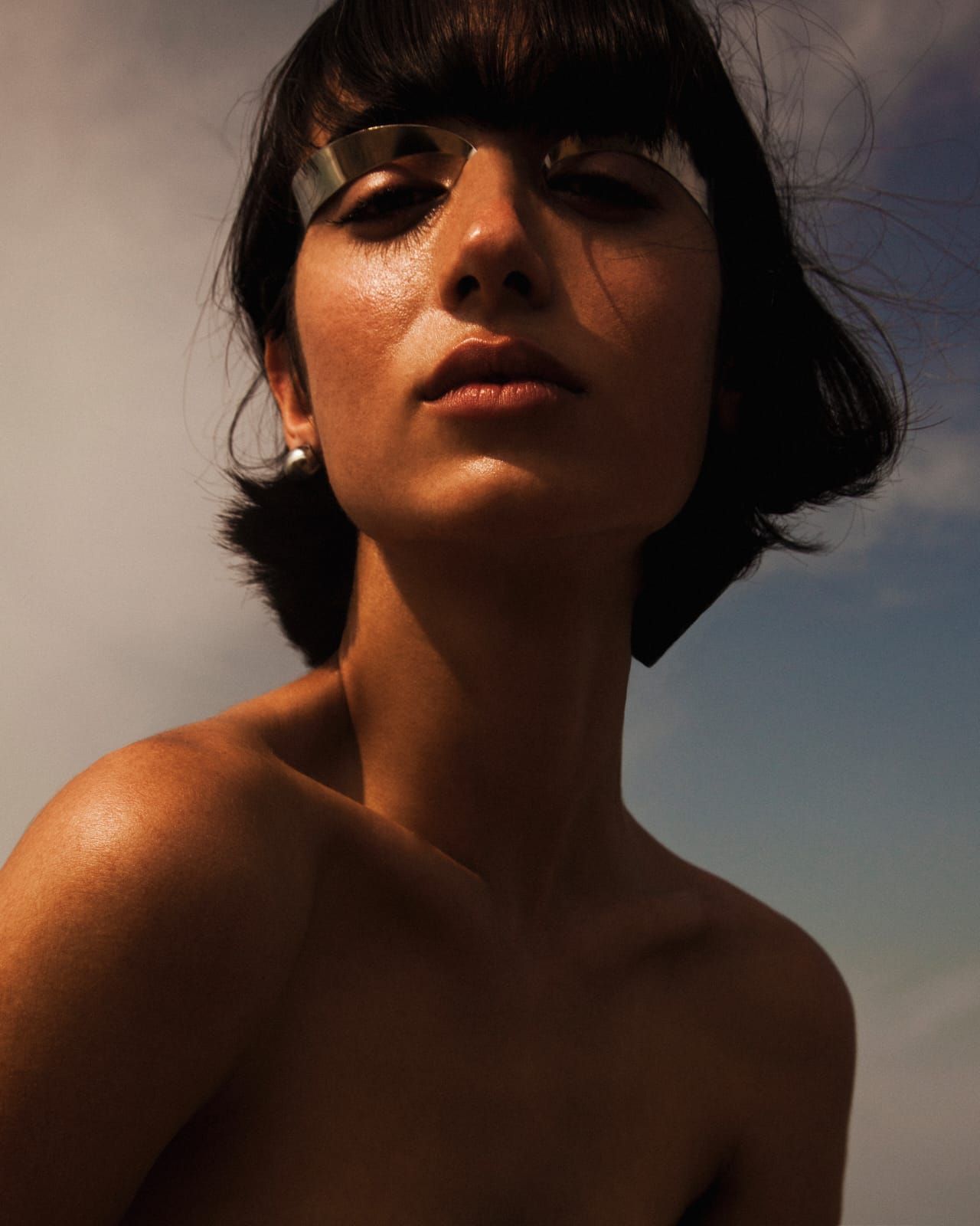 Woman with dark bob, metallic eye covers, and slightly damp skin, looking up with a neutral expression against a blue sky.