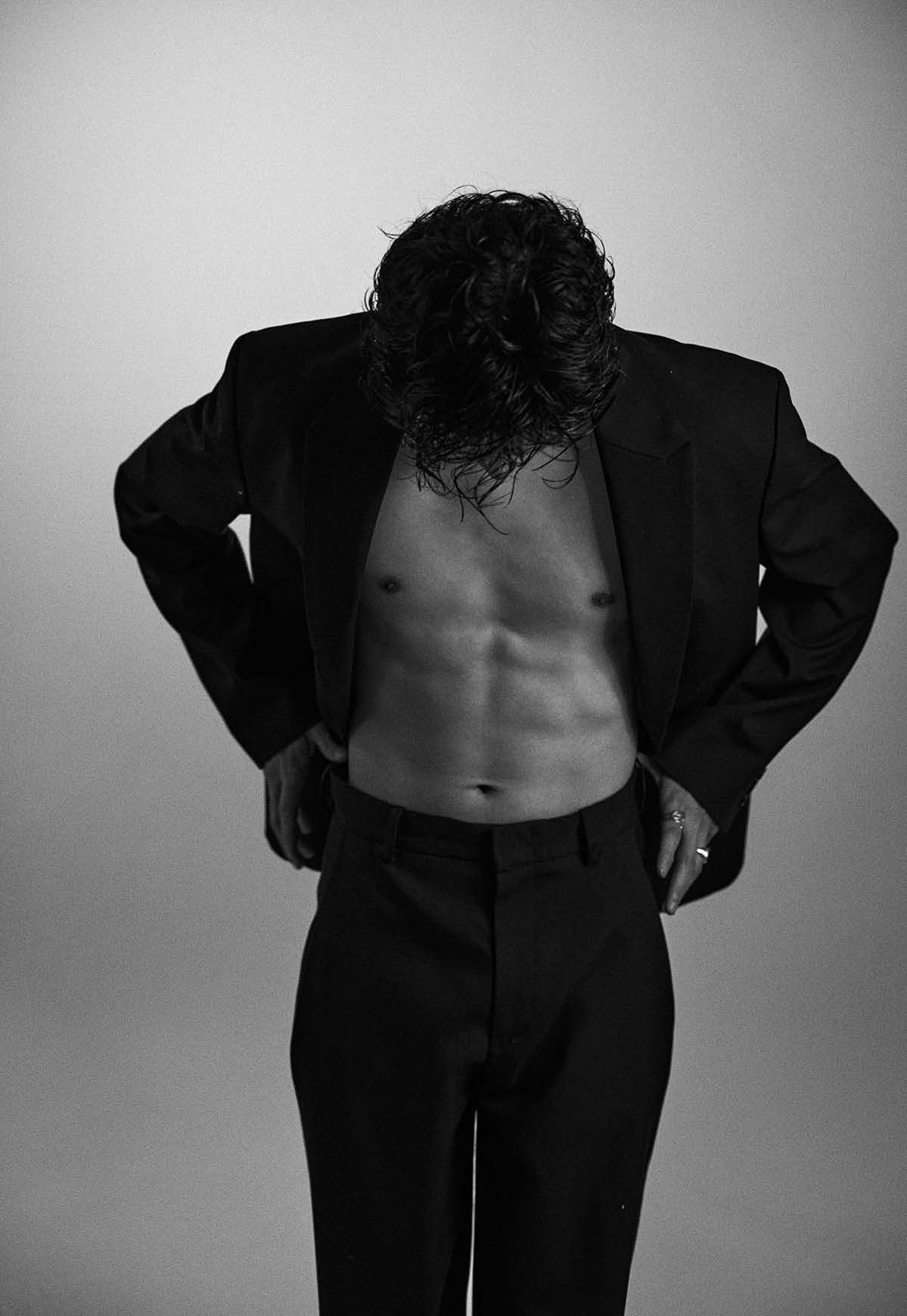 Man in a suit, unbuttoned, revealing defined abdominal muscles; leaning forward.