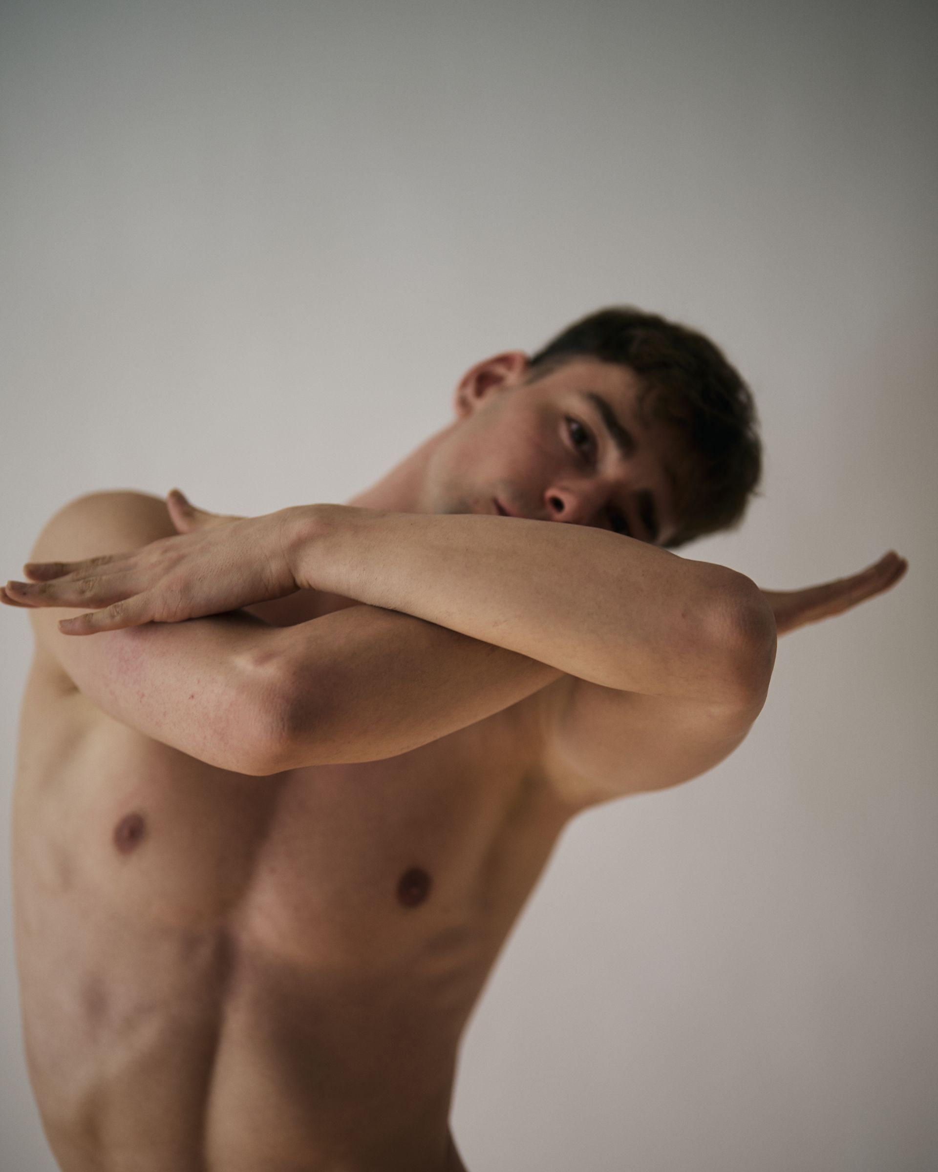Man, shirtless, arms crossed in front of face, looking towards the camera. Against a white background.