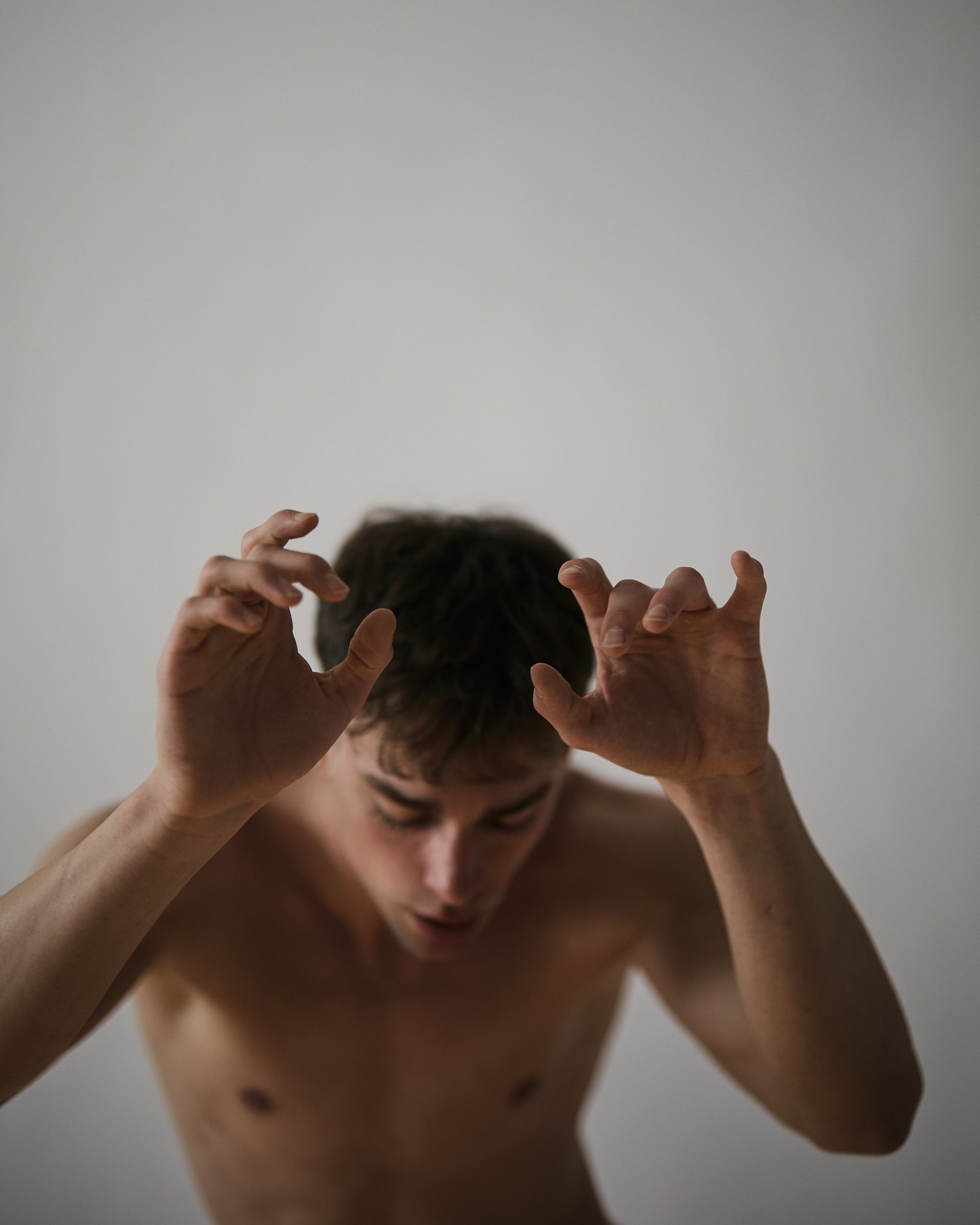 Man with hands raised, looking down. Bare chest. Neutral background.