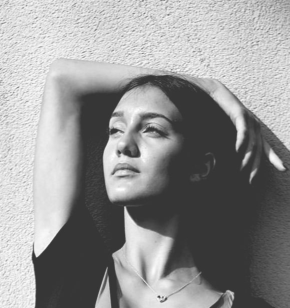 Woman in sunlight, arm raised, looking up, against a textured wall.