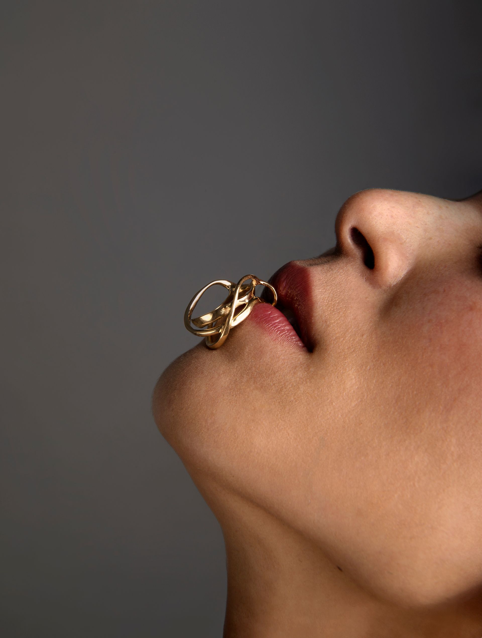 Close-up of a person's face with a gold ring in their mouth, lips parted, against a gray background.