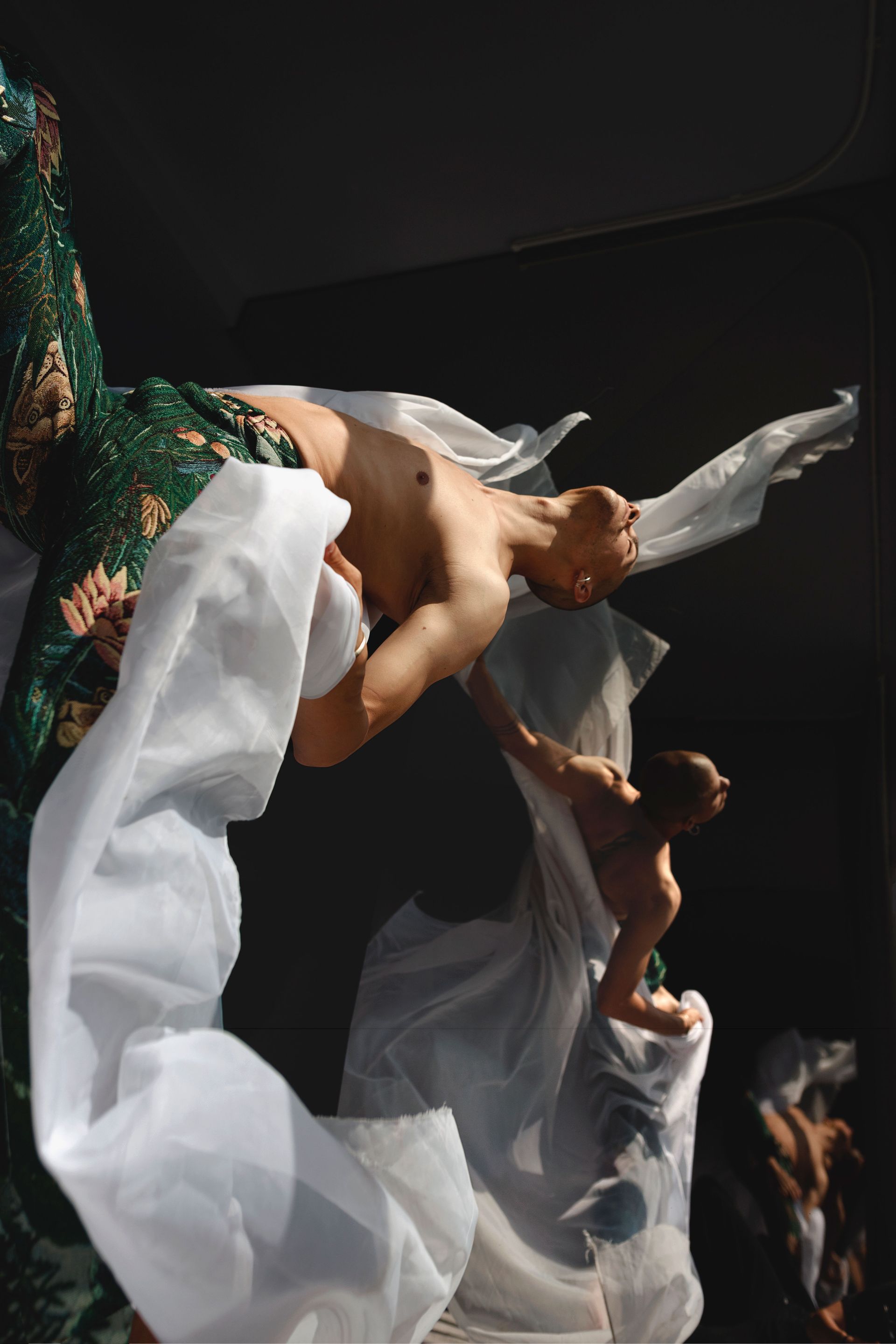 A person in a dance performance. They are shirtless and interacting with white fabric. Dark background.