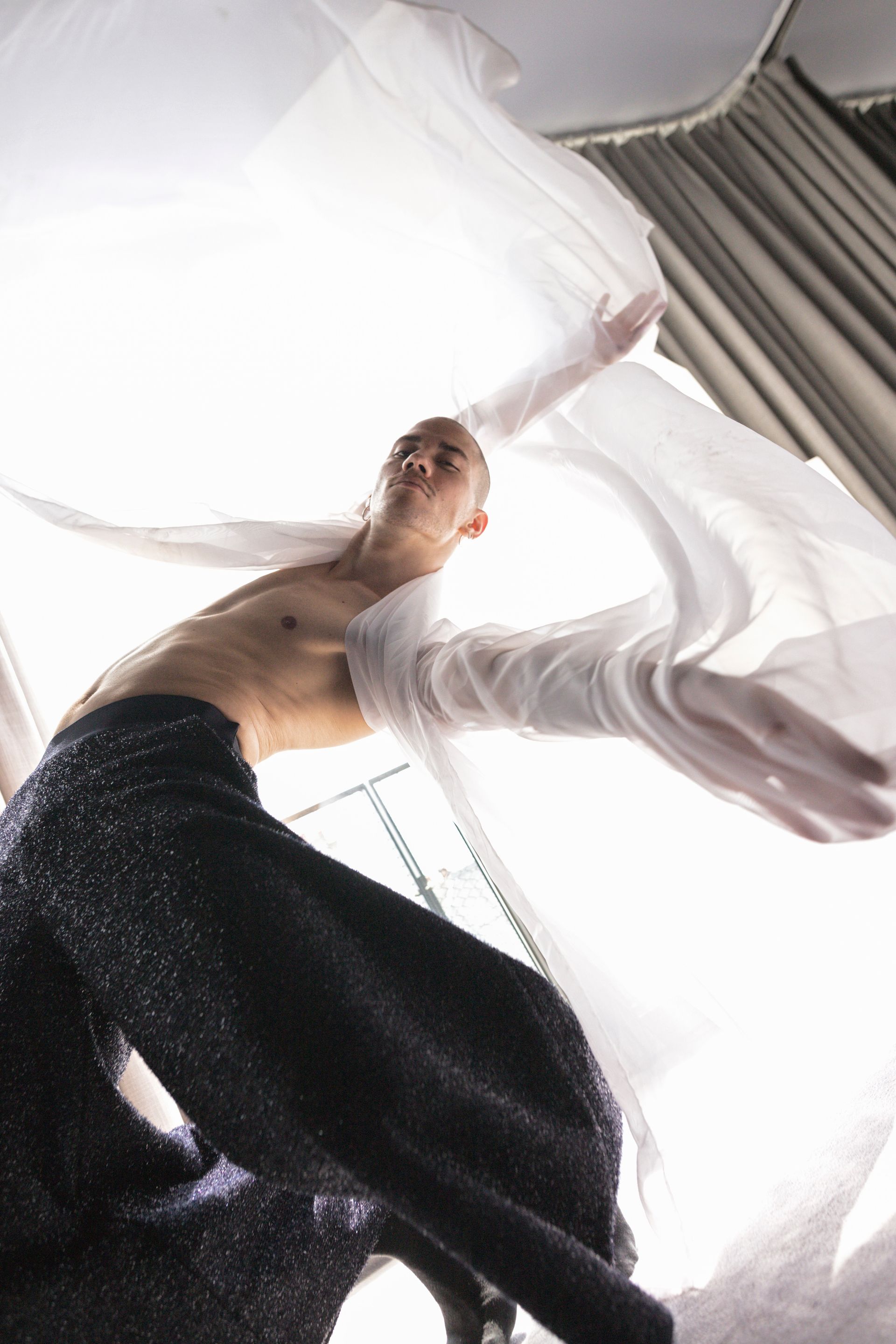 Man in flowing fabric, arms outstretched, wearing dark pants, body angled up with bright light.