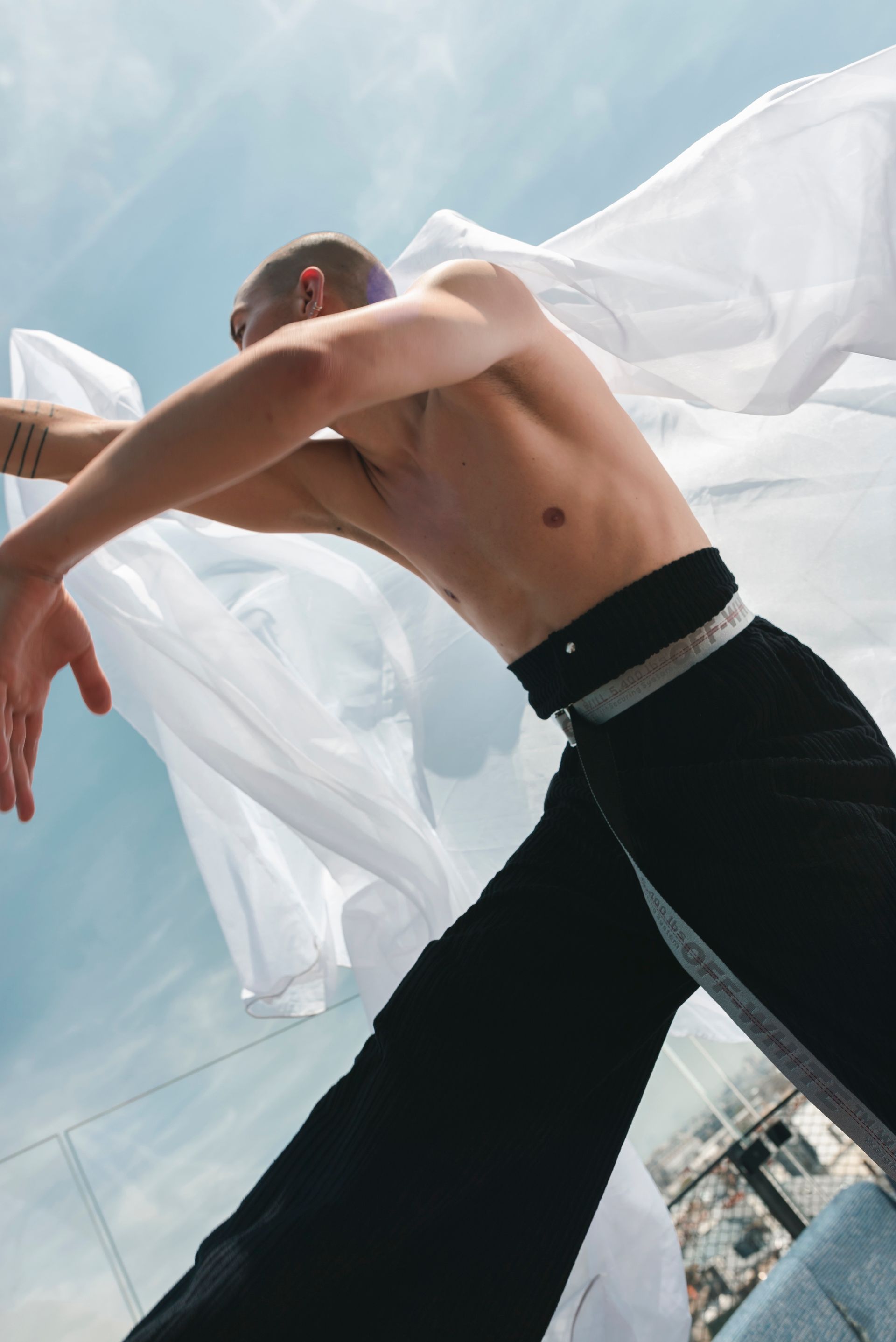 Man dancing with white fabric in front of a blue sky. He wears black pants.