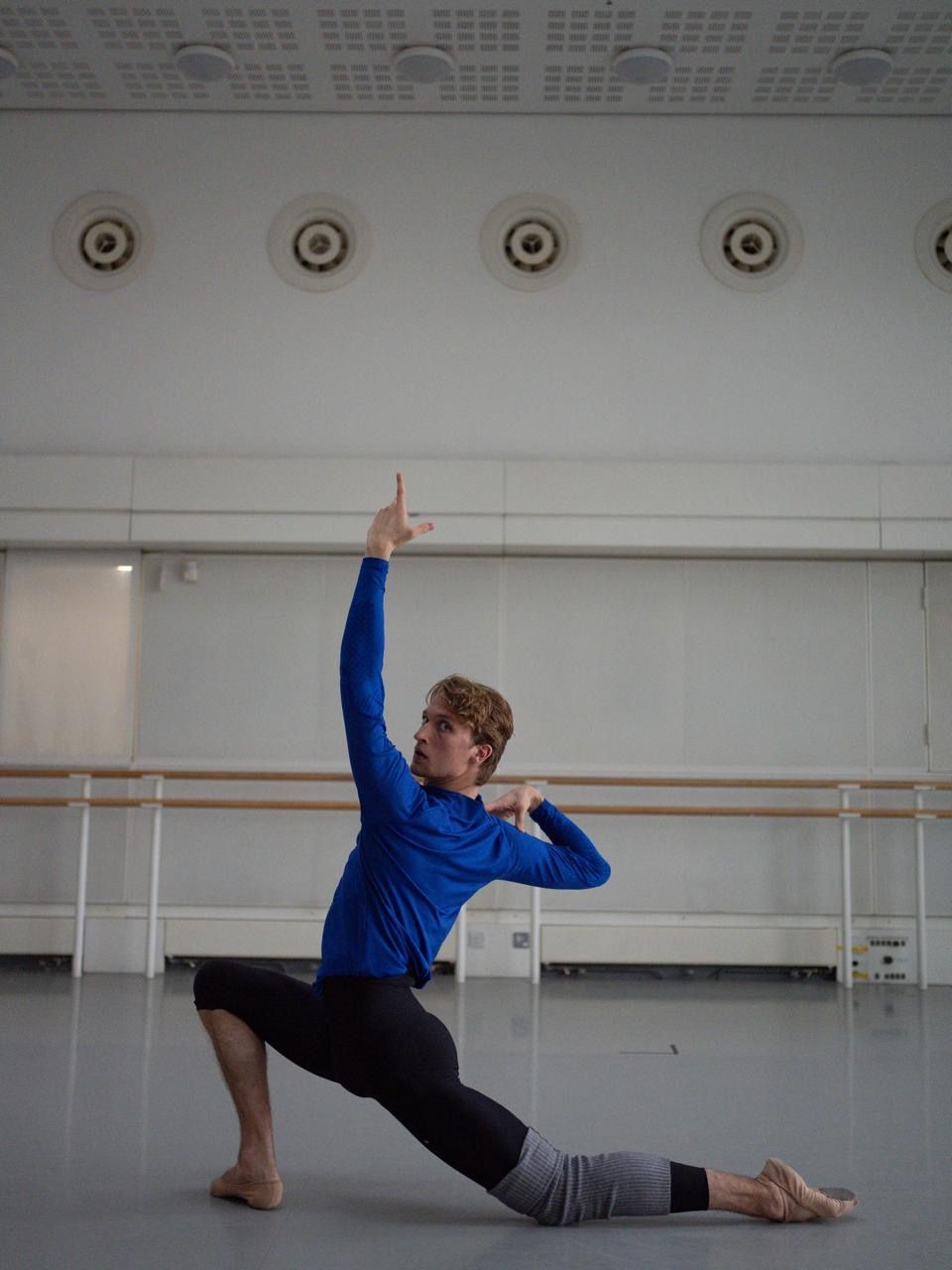 Ballet dancer in a deep lunge with one arm raised. Studio with barre. Blue shirt, black pants.
